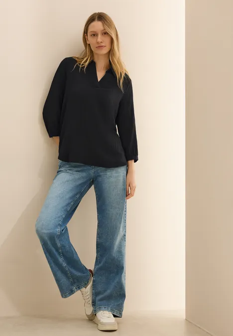 Frau in dunkler Bluse und Jeans steht lässig an einer Wand.